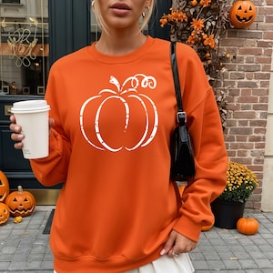 May include: An orange sweatshirt with a white pumpkin graphic. The long-sleeved top is a solid color with a distressed-style pumpkin design on the front. The image also shows a black purse, a white coffee cup, and decorative pumpkins.