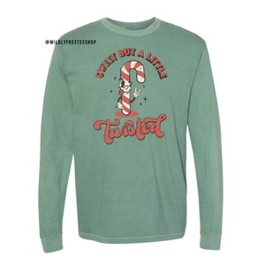 May include: A green long-sleeved t-shirt with a vintage-style graphic of a candy cane with a face and the text "Sweet but a little twisted".