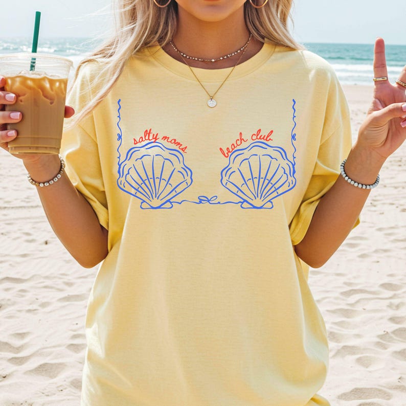 May include: A light yellow t-shirt with a blue seashell graphic and the text "salty moms beach club" in red and blue. The shirt is made of a soft material and is perfect for a day at the beach.