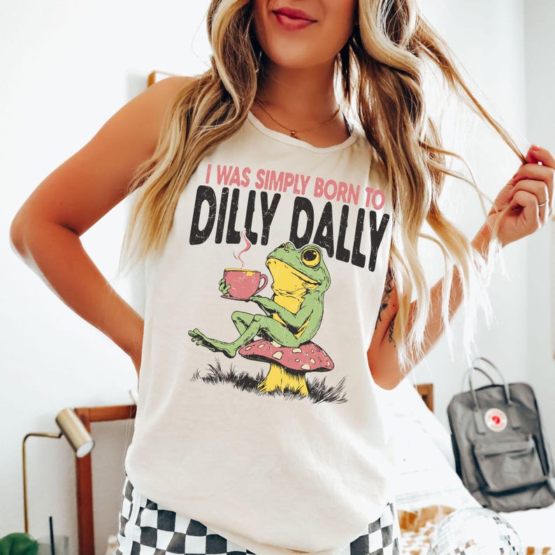 Op de afbeelding: Cr&egrave;mekleurige tanktop met de tekst "I WAS SIMPLY BORN TO DILLY DALLY." Een cartoonkikker zit op een paddenstoel en houdt een roze kopje vast. De kikker is groen en geel.