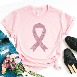 May include: A light pink short-sleeved t-shirt featuring a large, central awareness ribbon graphic with a textured, glittery pink finish. The garment has a crew neckline and a standard fit, shown laid flat with a label visible at the inner collar.