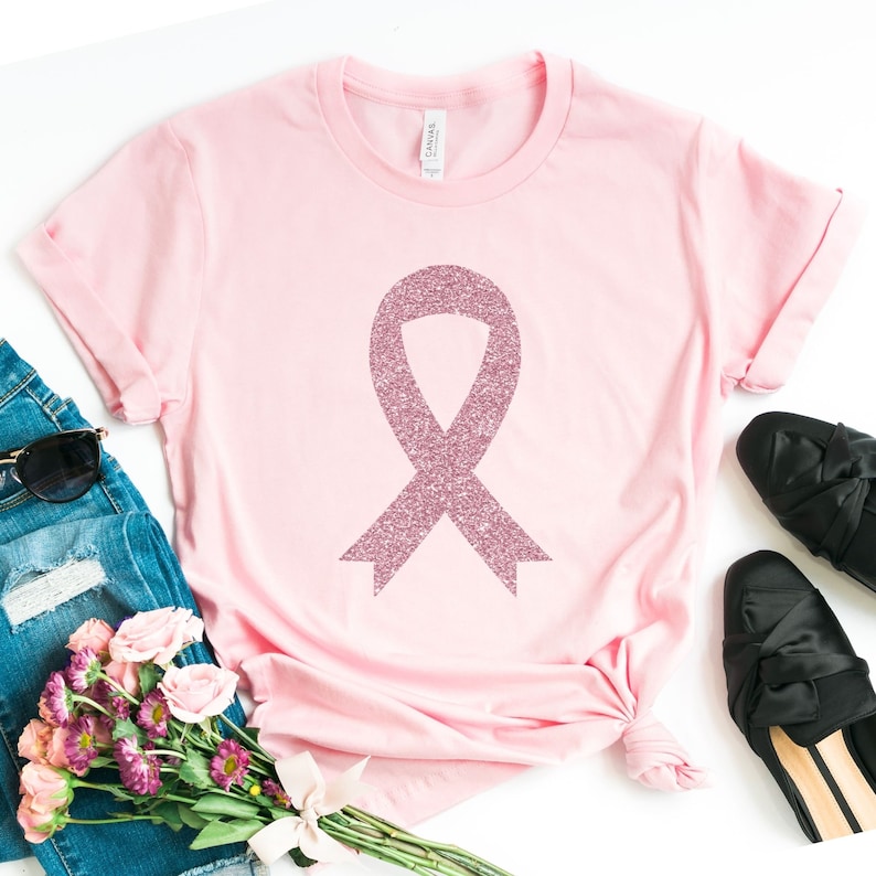 May include: A light pink short-sleeved t-shirt featuring a large, central awareness ribbon graphic with a textured, glittery pink finish. The garment has a crew neckline and a standard fit, shown laid flat with a label visible at the inner collar.