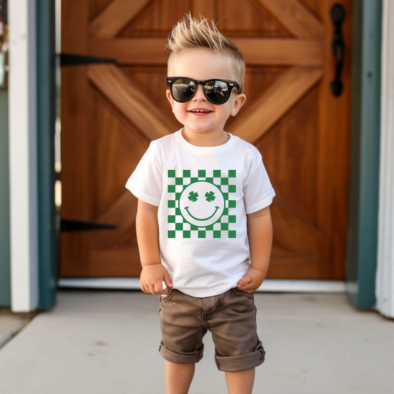 May include: A white t-shirt with a green and white checkered pattern and a smiley face with shamrock eyes. The shirt is worn by a young child wearing sunglasses and brown shorts.