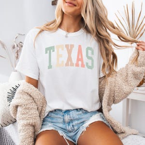 May include: White t-shirt with a colorful "TEXAS" graphic in a retro style. The shirt is worn by a person with long blonde hair and is paired with denim shorts.