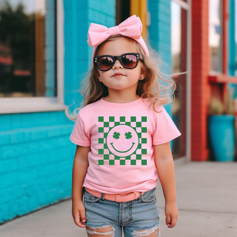May include: A pink t-shirt with a green and white checkered pattern featuring a smiley face with shamrock eyes. The child is wearing sunglasses and a pink bow. The shirt is paired with denim shorts and a pink belt.