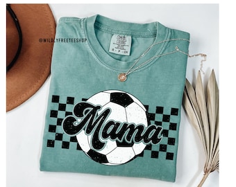 May include: A teal blue t-shirt with a black and white soccer ball graphic and the word "Mama" in a retro font. The graphic is surrounded by a black and white checkered pattern.