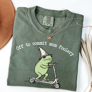 Puede incluir: Camiseta verde oliva con el texto "Off to commit mom foolery". La camiseta presenta una rana de dibujos animados con un sombrero de mago montada en un patinete. Un collar dorado con un colgante está sobre la camiseta.