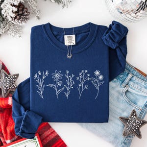 May include: A navy blue long-sleeve shirt with a white floral design. The shirt has a crew neck and is made of a soft material. A silver necklace with a sun pendant is on the shirt.