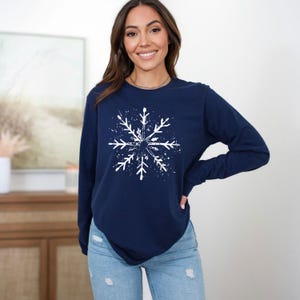 Op de afbeelding: Marineblauwe longsleeve met een wit sneeuwvlokontwerp. De sneeuwvlok is gecentreerd en heeft een spatteffect. De top wordt gecombineerd met een lichtblauwe jeans.