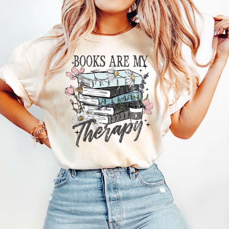May include: Cream-colored t-shirt with a graphic that reads "BOOKS ARE MY Therapy." The design features a stack of books, flowers, and a coffee cup. The books are in shades of blue, black, and white, with pink and white flowers surrounding them.