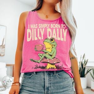 Op de afbeelding: Felroze tanktop met de tekst "I WAS SIMPLY BORN TO DILLY DALLY" en een afbeelding van een groene en gele kikker die op een paddenstoel zit en een kopje thee vasthoudt.
