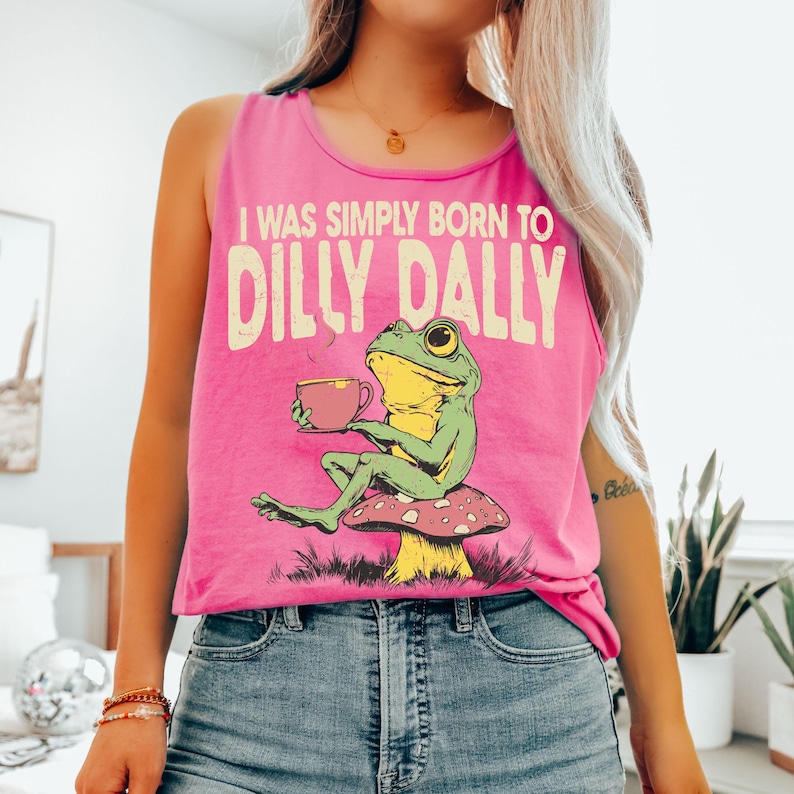 Op de afbeelding: Felroze tanktop met de tekst "I WAS SIMPLY BORN TO DILLY DALLY" en een afbeelding van een groene en gele kikker die op een paddenstoel zit en een kopje thee vasthoudt.
