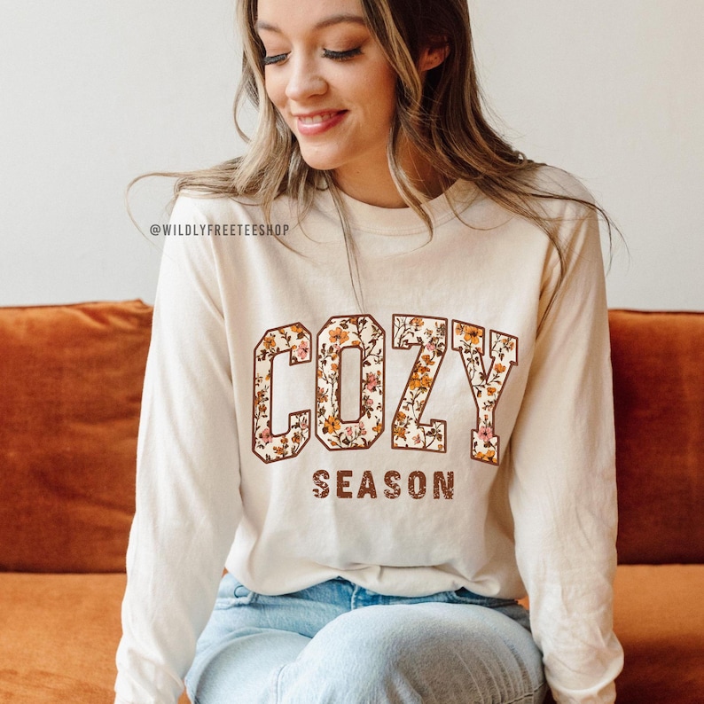 May include: Cream-colored long-sleeve shirt with the words "COZY SEASON" in a floral design. The shirt is made of a soft material and is perfect for fall. The shop's name, @WILDLYFREETEESHOP, is printed at the top.