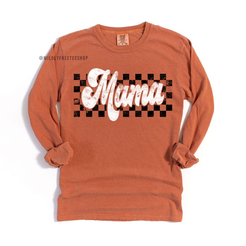 Puede incluir: Una camiseta de manga larga de color naranja quemado con un gr&aacute;fico blanco de "Mama" sobre un fondo a cuadros blanco y negro.