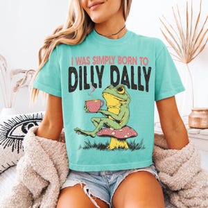 May include: Teal t-shirt featuring the text "I WAS SIMPLY BORN TO DILLY DALLY" and a cartoon frog holding a pink teacup, seated on a mushroom. The frog is green and yellow. The shirt has a relaxed fit.