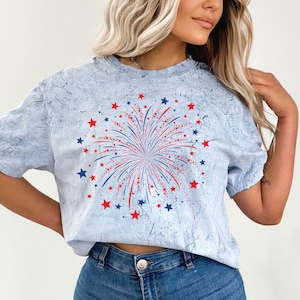 Op de afbeelding: Een lichtblauw T-shirt met een rood, wit en blauw vuurwerkmotief. Het vuurwerk is omringd door rode en blauwe sterren.