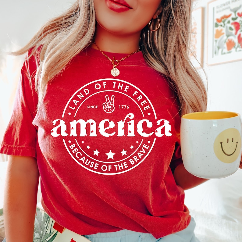 May include: Red t-shirt with a white graphic that reads "Land of the Free, America, Because of the Brave" with a peace sign and stars. A white mug with a yellow interior and a smiley face is held in the hand.