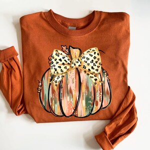 May include: An orange long-sleeved shirt featuring a pumpkin design. The pumpkin has a bow with a leopard print pattern. The pumpkin design has a mix of colors including orange, pink, green, and gold. The sleeves are rolled up.