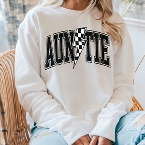 K&ouml;nnte beinhalten: Ein wei&szlig;es lang&auml;rmeliges Sweatshirt mit Rundhalsausschnitt und dem schwarzen Schriftzug AUNTIE auf der Brust. Der Buchstabe I ist durch einen schwarz-wei&szlig; karierten Blitz ersetzt. Die Schrift hat eine abgenutzte, verwitterte Textur.