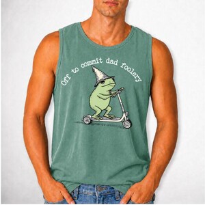 Könnte beinhalten: Ein salbeigrünes ärmelloses Tanktop mit einer grafischen Illustration eines hellgrünen Frosches, der einen spitzen Hut trägt und auf einem Tretroller fährt. Der Text "Off to commit dad foolery" ist in Weiß in einem Bogen über dem Frosch gedruckt.