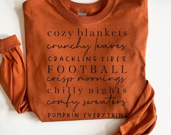 May include: An orange long-sleeved shirt with black text listing fall-themed phrases: "cozy blankets," "crunchy leaves," "crackling fires," "football," "crisp mornings," "chilly nights," "comfy sweaters," and "pumpkin everything."