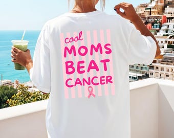 May include: A white t-shirt featuring a back graphic with vertical light pink stripes and the text "cool MOMS BEAT CANCER" in bold pink lettering, accompanied by a small pink awareness ribbon icon positioned below the text.