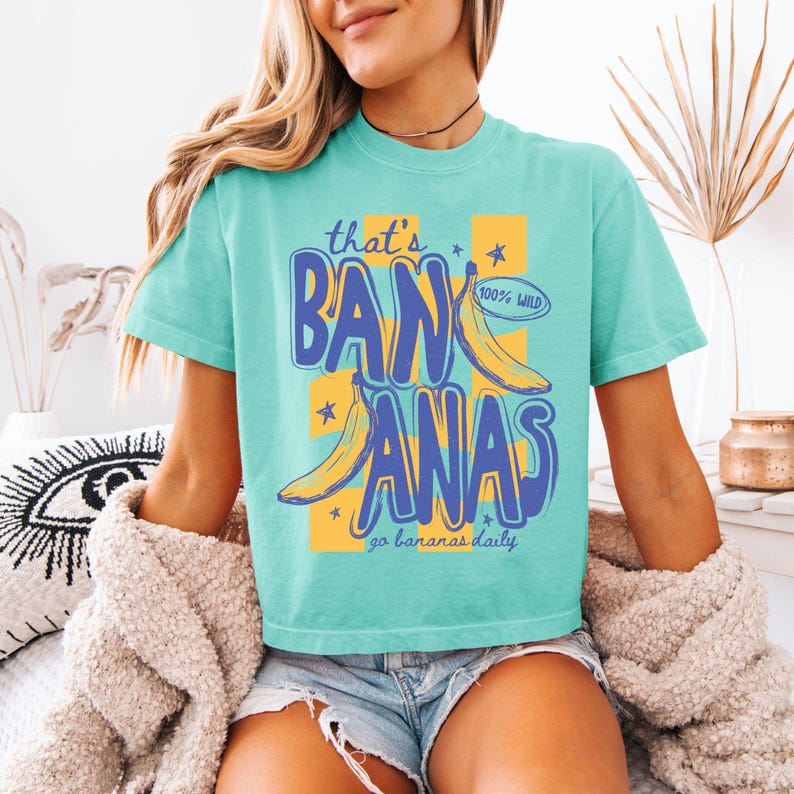 Pu&ograve; includere: Una maglietta turchese con la scritta "that's BANANAS" in blu e giallo, con illustrazioni di banane e il testo "100% WILD" e "go bananas daily".