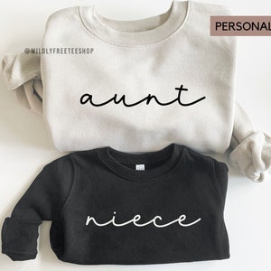May include: Two sweatshirts, one beige and one black, with the words "aunt" and "niece" written in cursive on them. The sweatshirts are personalized and can be customized with different names.