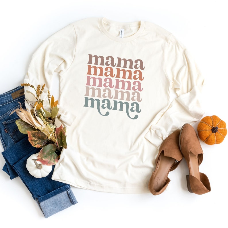 May include: A long-sleeved, cream-colored t-shirt with the word 'mama' repeated five times in a stacked design. The word 'mama' is in a retro font and is colored in shades of pink, orange, and blue.
