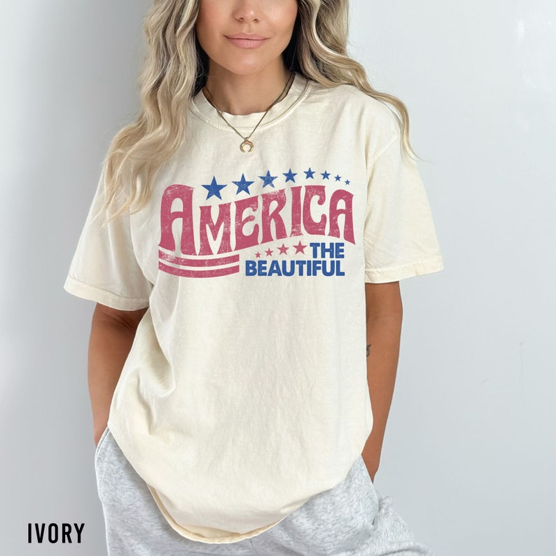 Pu&ograve; includere: Una t-shirt color crema con una grafica in stile vintage che dice "America The Beautiful" in lettere rosse e blu con stelle.