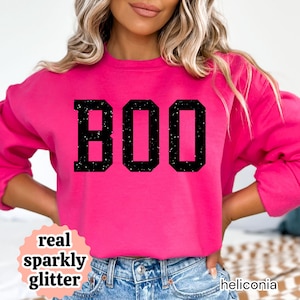 May include: A bright pink crewneck sweatshirt featuring the word BOO printed in large, black, block-style letters with a speckled, glittery texture. The garment has long sleeves and a ribbed neckline, providing a simple and casual appearance.