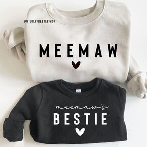 Könnte beinhalten: Zwei Sweatshirts, eines in Hellbeige und eines in Schwarz. Das beige Sweatshirt hat das Wort "MEEMAW" in schwarzen Blockbuchstaben mit einem schwarzen Herz darunter. Das schwarze Sweatshirt sagt "meemaw's BESTIE" in weißer Schrift und Blockbuchstaben mit einem weißen Herz.