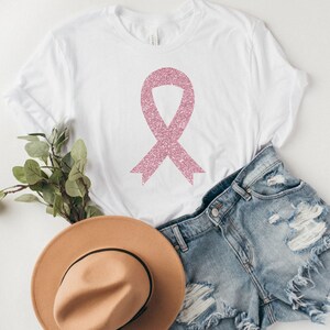 May include: A white short-sleeved t-shirt featuring a pink glittery awareness ribbon graphic centred on the chest. The shirt is displayed alongside a pair of distressed blue denim shorts and a tan wide-brimmed hat with a black band.