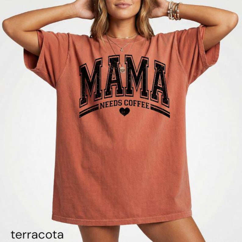 May include: A terracotta-coloured short-sleeve t-shirt featuring black distressed lettering that reads MAMA NEEDS COFFEE with a small heart icon centred below the text. The shirt has a relaxed fit and a crew neckline, showcasing a vintage-inspired graphic design printed on the front.