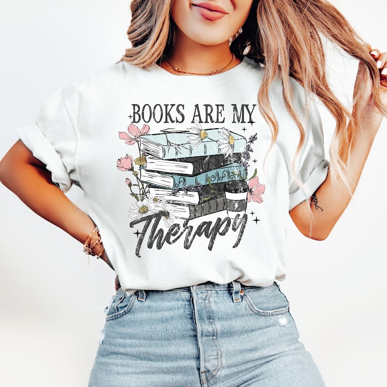 May include: White t-shirt with a graphic of stacked books, flowers, and the text "BOOKS ARE MY Therapy". The design features a coffee cup and decorative floral elements. The shirt is made of a soft material.