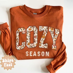 May include: An orange long-sleeved sweatshirt with the words "COZY SEASON" in a floral design. The word "COZY" is in large, block letters with a floral pattern, and "SEASON" is in a smaller font below. The sleeves are cuffed.