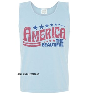 Pu&ograve; includere: Una canotta azzurra con una grafica rossa e blu che dice "America The Beautiful" con stelle intorno alla parola "America".