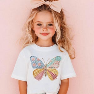以下が含まれることがあります： カラフルな蝶のデザインが施された白いTシャツ。蝶は、青、ピンク、黄色、赤の色合いの花柄、チェック柄、無地のパッチワーク生地で作られています。金髪の幼い子供がそのTシャツを着ています。