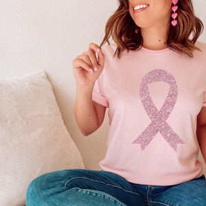 May include: A light pink short-sleeved t-shirt featuring a large, centred awareness ribbon graphic. The ribbon design is rendered in a textured, sparkling mauve glitter finish. The shirt has a classic crew neckline and cuffed sleeves.