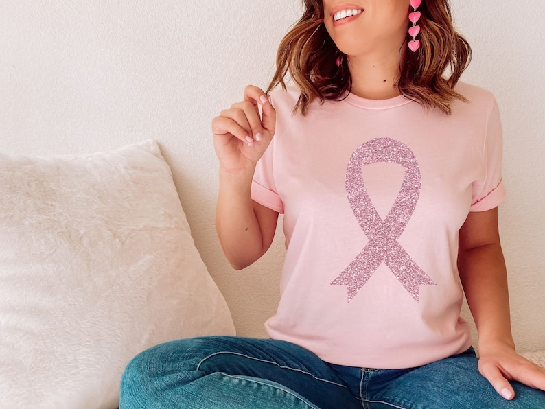 May include: A light pink short-sleeved t-shirt featuring a large, centred awareness ribbon graphic. The ribbon design is rendered in a textured, sparkling mauve glitter finish. The shirt has a classic crew neckline and cuffed sleeves.
