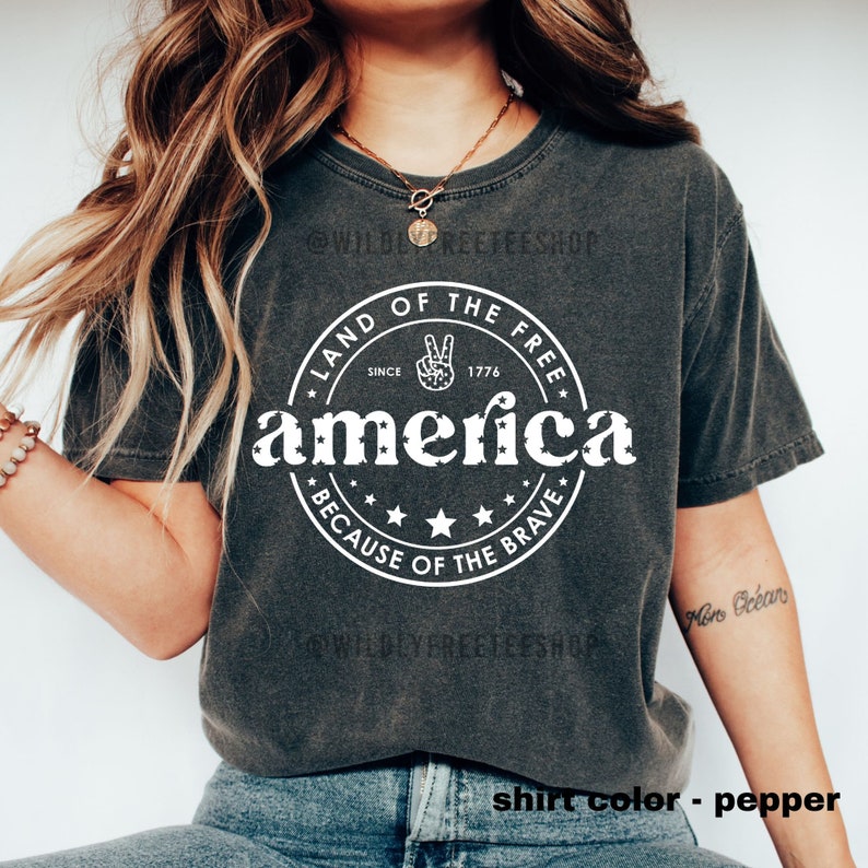 May include: Pepper-colored t-shirt with a white circular graphic that reads "Land of the Free" and "America" with stars and the text "Because of the Brave." The graphic includes a peace sign and the year 1776.