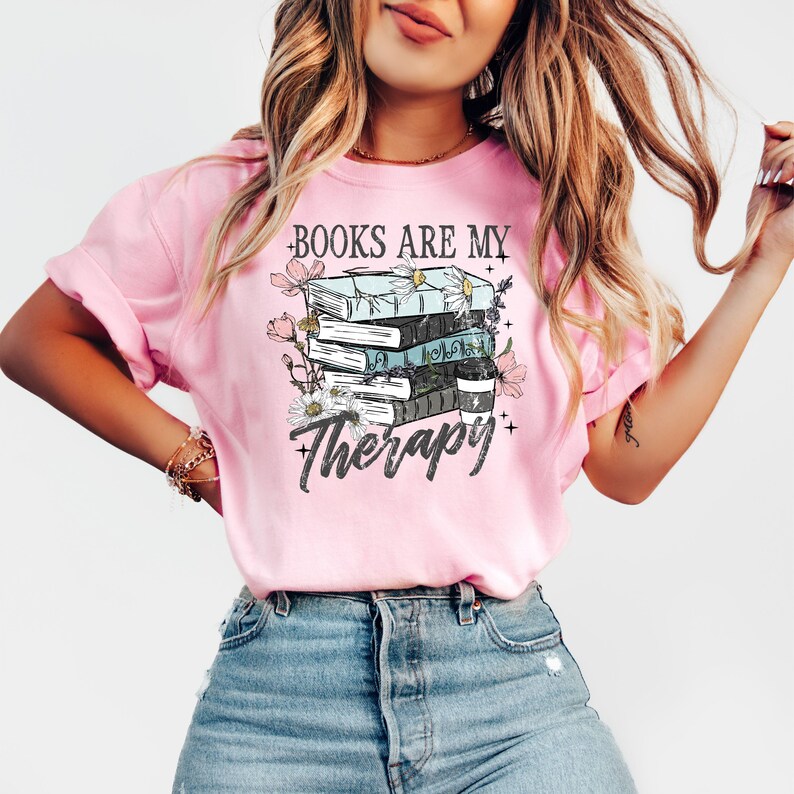 May include: Pink t-shirt with the text "BOOKS ARE MY Therapy" and a graphic of stacked books, flowers, and stars. The books are in shades of blue, black, and white. The shirt has short sleeves and a crew neck.
