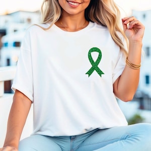 May include: A white t-shirt with a green glitter ribbon on the left chest. The ribbon is a symbol of awareness for various causes.