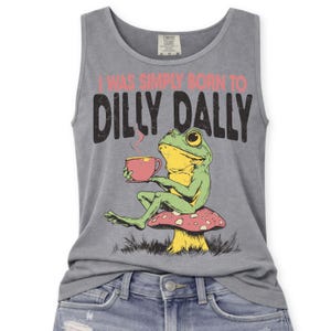 Op de afbeelding: Grijze tanktop met de tekst "I WAS SIMPLY BORN TO DILLY DALLY" en een cartoon kikker met een theekopje. De kikker zit op een paddenstoel. De tanktop is gemaakt van een zacht materiaal.