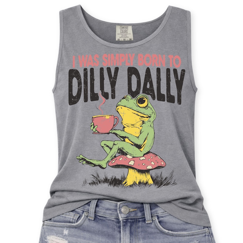 Op de afbeelding: Grijze tanktop met de tekst "I WAS SIMPLY BORN TO DILLY DALLY" en een cartoon kikker met een theekopje. De kikker zit op een paddenstoel. De tanktop is gemaakt van een zacht materiaal.