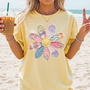 Peut inclure: T-shirt jaune clair orné d'une grande fleur colorée. La fleur est composée de différents patchworks de tissus aux tons roses, bleus et jaunes. Le t-shirt a des manches courtes et une coupe décontractée, idéal pour un look décontracté.