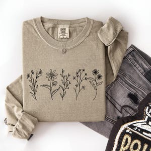May include: A tan long-sleeve shirt with a crew neck and a black floral line art design across the front. A silver necklace with a sun pendant is on the shirt. The shirt is folded with a pair of gray jeans.