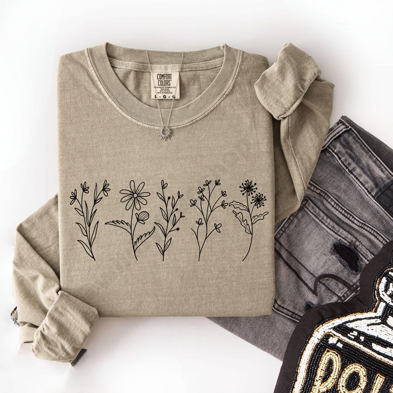May include: A tan long-sleeve shirt with a crew neck and a black floral line art design across the front. A silver necklace with a sun pendant is on the shirt. The shirt is folded with a pair of gray jeans.