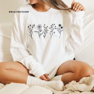 May include: White long-sleeve shirt featuring a black floral line art design across the chest. The shirt has a crew neck and long sleeves. The text "@WILDLYFREETEESHOP" is printed at the top left of the shirt.
