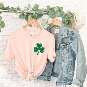 May include: A pink t-shirt with a green glitter shamrock design. The shirt is hanging on a rack with a denim jacket and a straw hat.